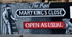 Zu sehen ist der Schriftzug "The Real Mary King's Close", sowie ein "open Asusual". Am rechten unteren Rand ist eine graue Ratte zu sehen und links ein Mann mit einer Schnabelmaske, wie wir gegen die Pest benutzt wurden.