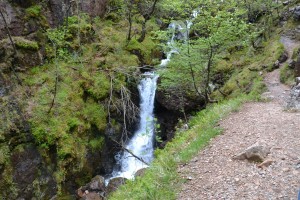In der Mitte des Bildes ist ein Wasserfall zu sehen, der einige Meter herunter fällt. Im rechten Teil ist der Weg zu sehen, neben dem ein Abhang verläuft.